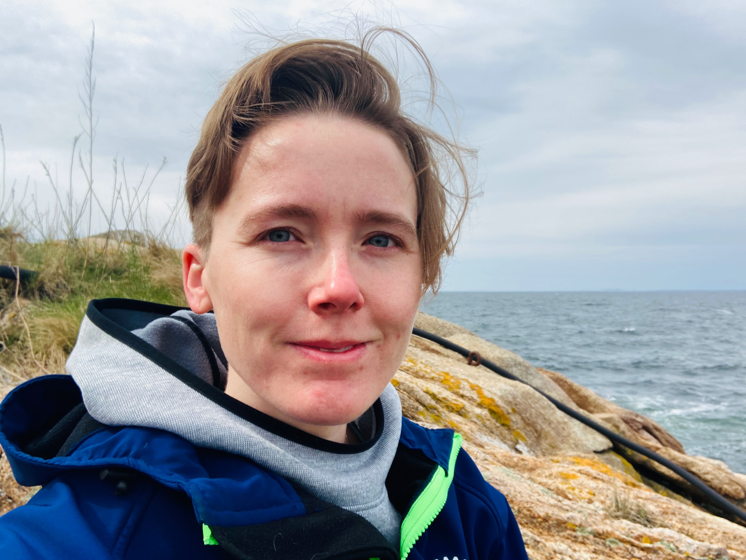 IMG_7339.jpeg Photo of a smiling person with short hair in front of rocks and ocean