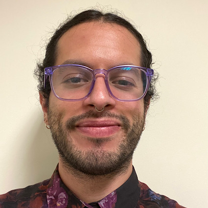 Eddie N..jpeg Non-binary person smiling wearing purple glasses.