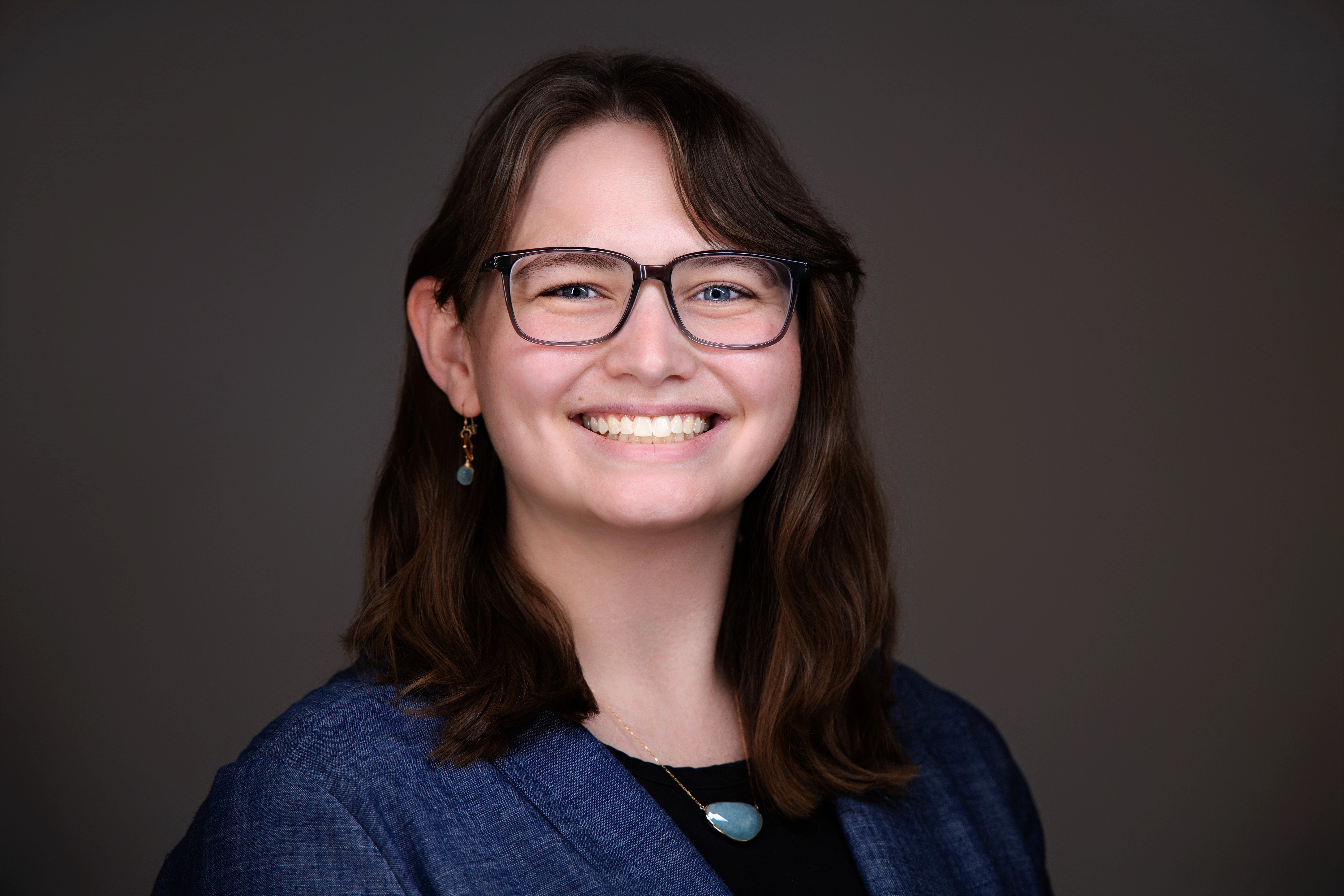 a young woman with brown hair and glasses smiling wearing a black dress and a blue blazer