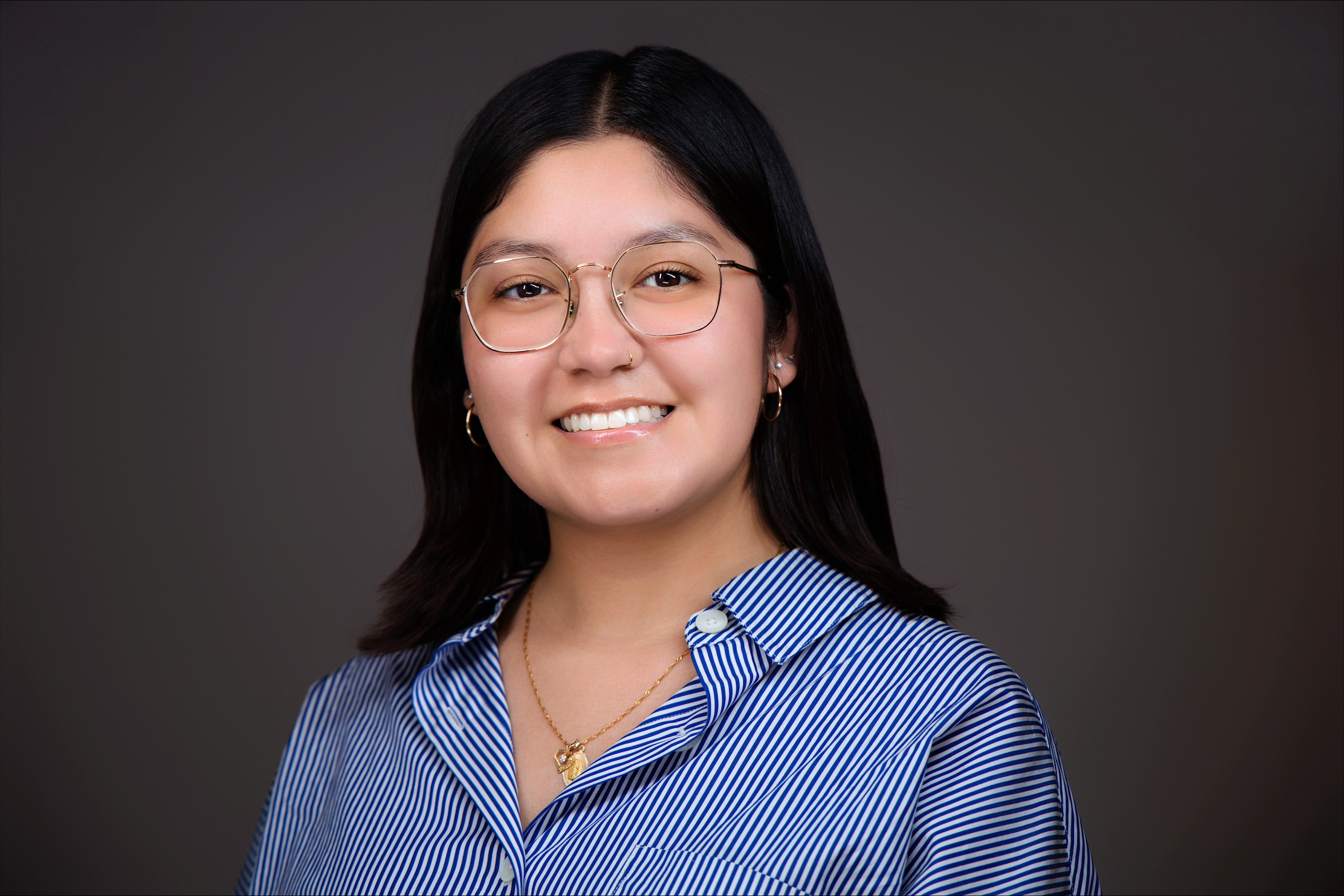Andrea Serna is pictured with a blue stripped button down, glasses, and straight dark hair.