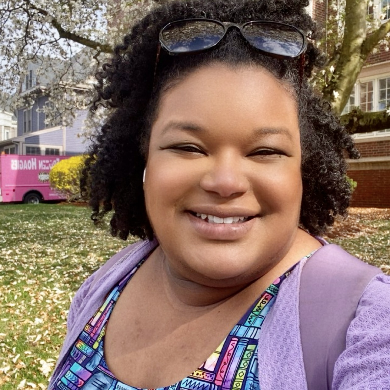 A brown-skinned woman with medium length, dark, curly hair smiles in front of a blooming tree. She is wearing a colorful book dress and a lilac cardigan and backpack.