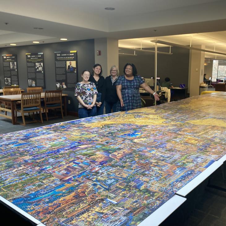 Four women smile as they stand behind a very large puzzle on display.