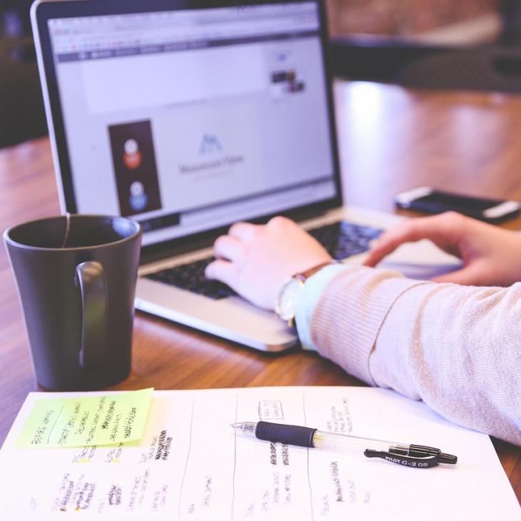 Person with only their arms visible working on a laptop, with mug &amp; paper &amp; pen