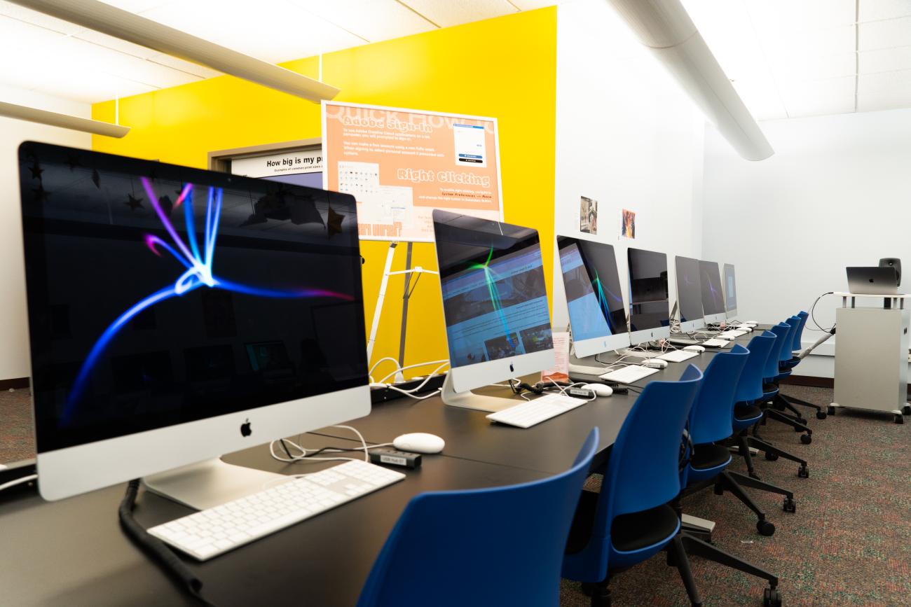 Row of various iMac's with yellow wall in background and rolling blue chairs in foreground