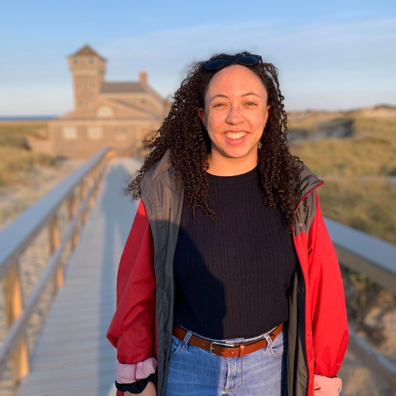 Portrait of Cana standing on a boardwalk. She has curly brown hair, is smiling and wearing a bright red coat, black shirt and jeans.