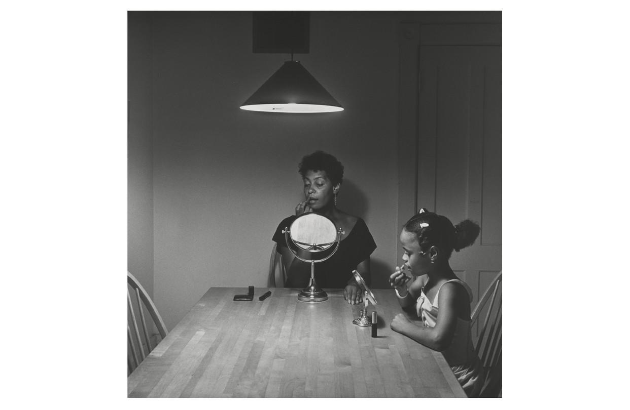 Black and white image of a black woman and child sitting at a table, both looking in individual mirrors while applying lipstick.