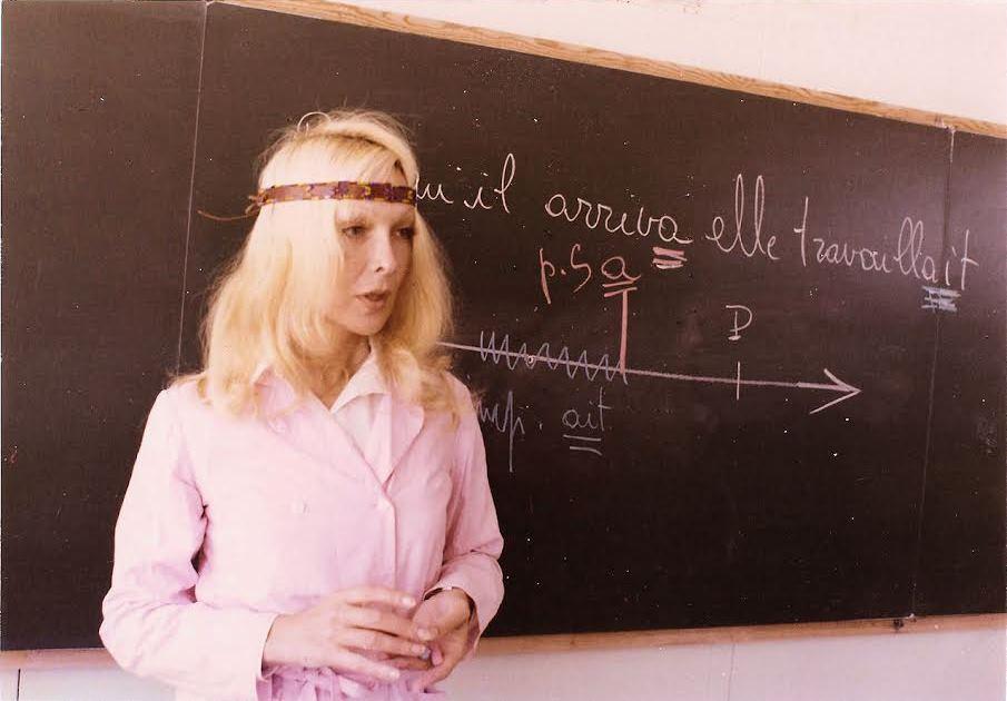 Person with long blonde hair standing in from of chaulk board.
