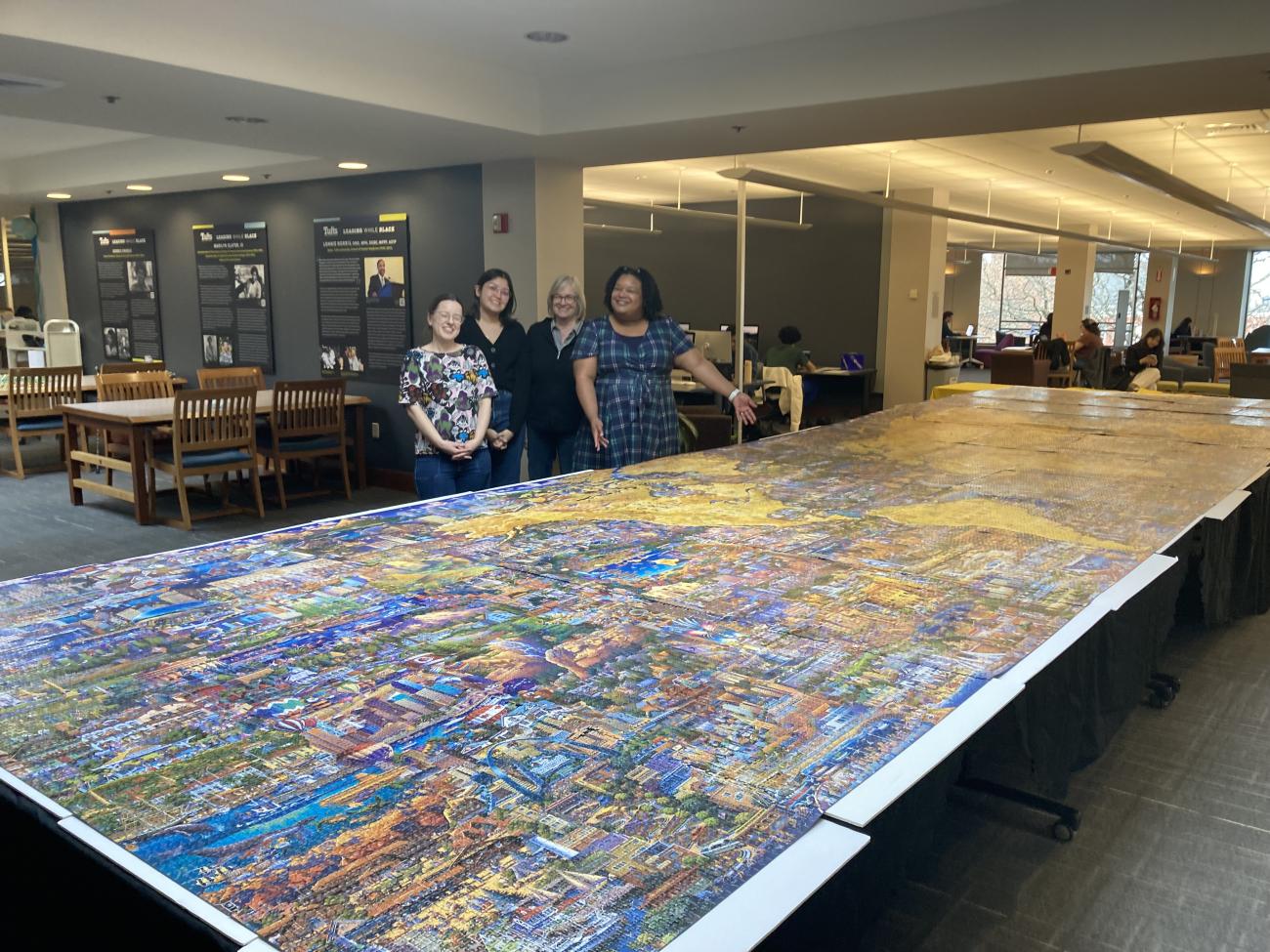 Four women smile as they stand behind a very large puzzle on display.