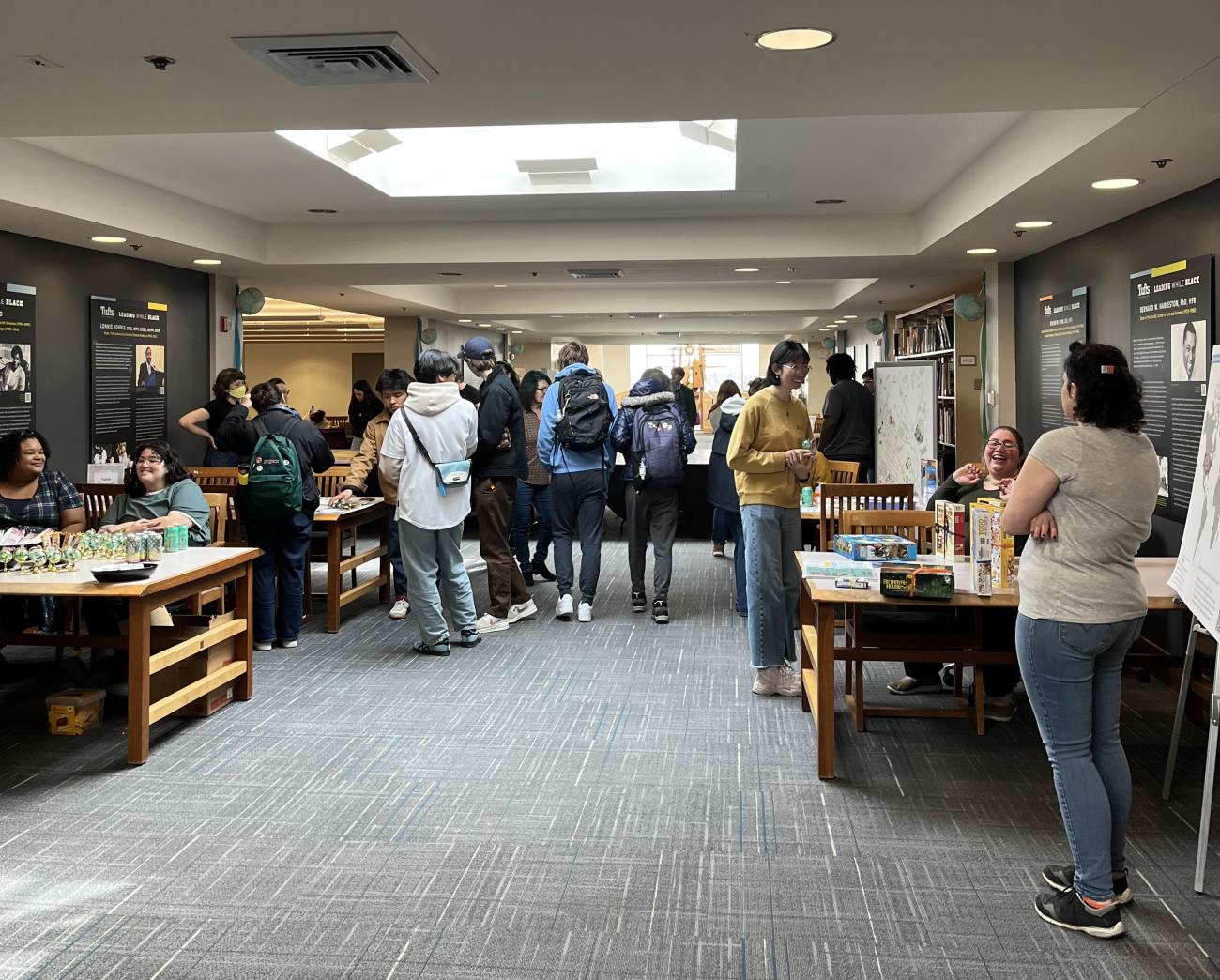 A crowd of students stand and walk through a corridor framed by library tables. Librarians and library staff sit and stand at each table with snacks and swag on display.