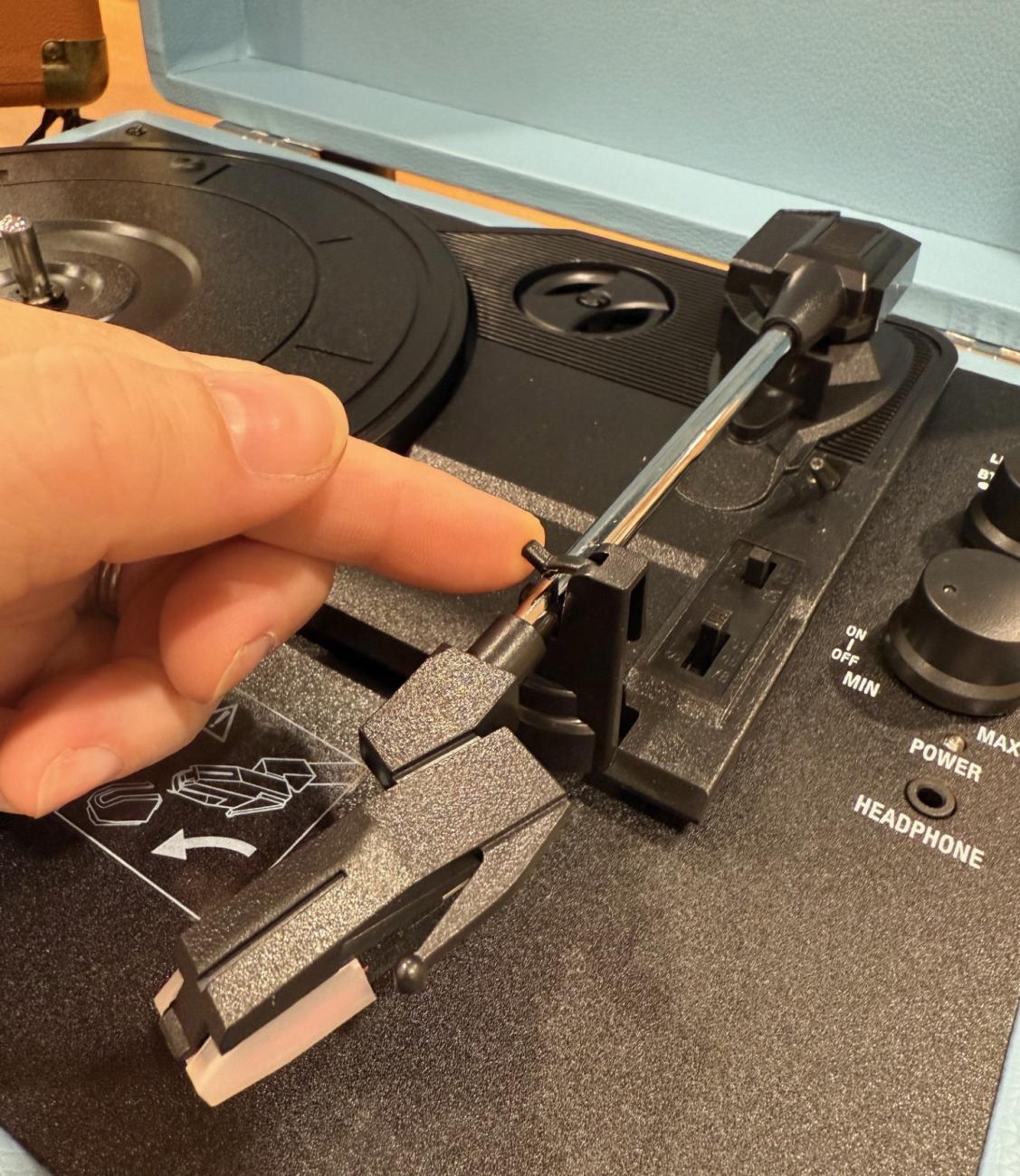a hand is showing the latch that holds the tonearm of the record player. It should be released to play a record, and replaced when not in use to keep the arm in place. .