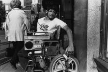 Black and white photograph of a main in a wheelchair with a 1980s style boombox with him.