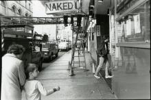 Black and white image of city sidewalk. Mother and child walking in the foreground towards a sign that reads &quot;Naked&quot;.  The childs gaze is towards the entrance of the building while the mothers gaze is down.