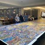 Four women smile as they stand behind a very large puzzle on display.