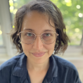 ForeroArnias2.png White skin person with short brown hair, wearing glasses and a blue top with green blurred foliage in background