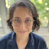 White skin person with short brown hair, wearing glasses and a blue top with green blurred foliage in background