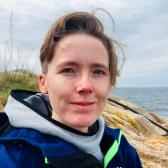 IMG_7339.jpeg Photo of a smiling person with short hair in front of rocks and ocean