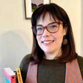 Smiling woman with light skin, medium brown hair, and red-purple eyeglasses stands in front of a small embroidered canvas holding three books cropped out of frame.