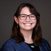 a young woman with brown hair and glasses smiling wearing a black dress and a blue blazer