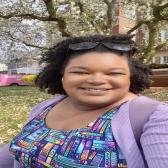 A medium brown-skinned woman with shoulder-length and dark curly hair in a half-updo smiles. She is wearing black sunglasses on her head, a colorful bookshelf dress, and a lilac cardigan.