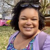 A brown-skinned woman with medium length, dark, curly hair smiles in front of a blooming tree. She is wearing a colorful book dress and a lilac cardigan and backpack.