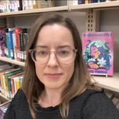 photo of Joanna wearing glasses and a black shirt, standing in front of a bookstack