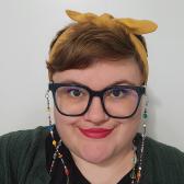 White individual wearing teal glasses and a rainbow glass bead eyeglass chain, a black shirt, green sweater, and yellow headband against a white background.